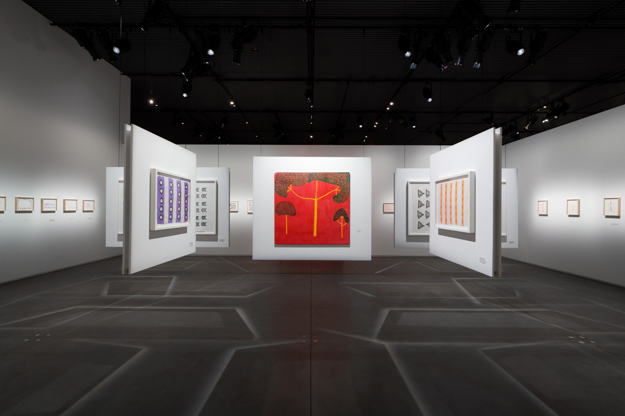 An art gallery with drawings and paintings on walls suspended from the ceiling and on fixed walls. The suspended walls make shadows of intersecting lines on the gray concrete floors. Situated prominently in the center of the gallery is a painting of three trees with yellow trunks and green canopies against a bright red background.