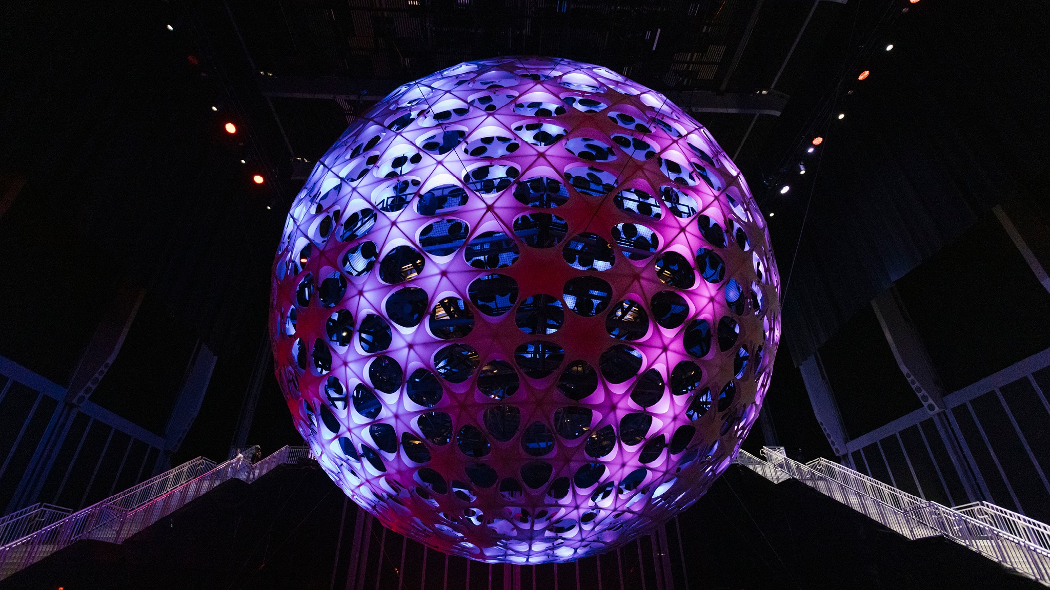 A vast spherical concert hall hangs from a 115-foot ceiling in a dark performance space. The sphere is dotted with nodes that light up in purple and blue light. Flanking the sphere are two sets of metal staircases that rise to the equator of the sphere.