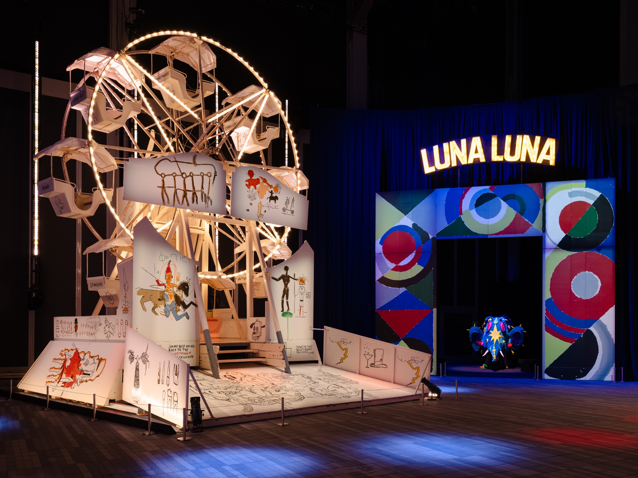 Two art carnival attractions sit side by side in a vast gallery exhibition space. One is a painted Ferris wheel by Jean-Michel Basquiat. It is painted an off-white and lit by bright white lights, with the artist's characteristic drawings and graffiti-inspired words painted on its surface. The second is an entry archway by Sonia Delaunay, painted in red, purple, blue, and green swirls with a lit sign above it announcing "Luna Luna."