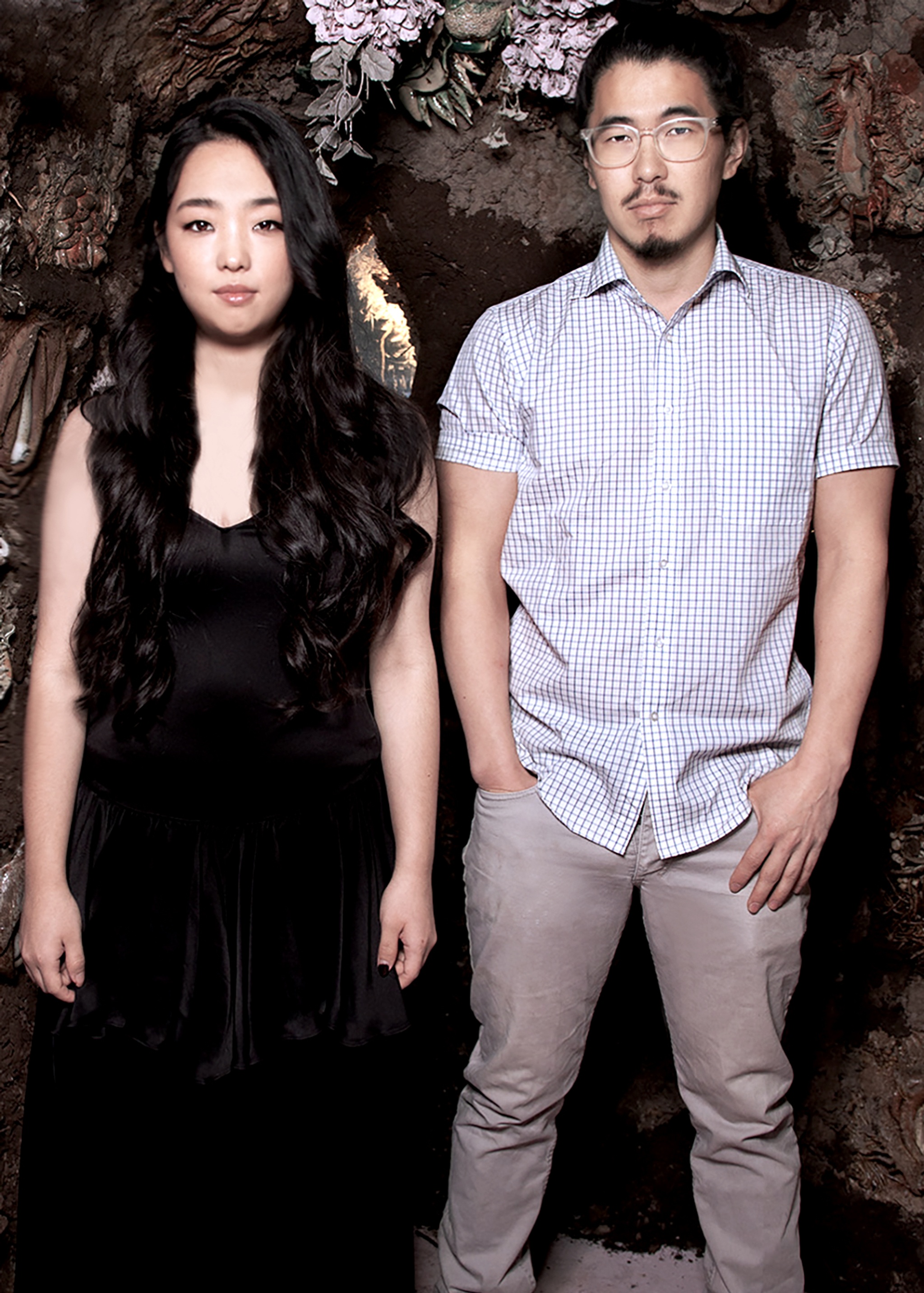 A portrait of artists A young Yu and Nick Oh. A young Yu is a Korean American woman and Nick is a Korean American man. They stand side by side, with shoulders touching and facing us. A young wears a long black dress and has long straight black hair that hangs beneath her shoulders. Nick has a mustache and goatee, wears glasses and a button down shirt, and stands with one hand in his pants pocket.