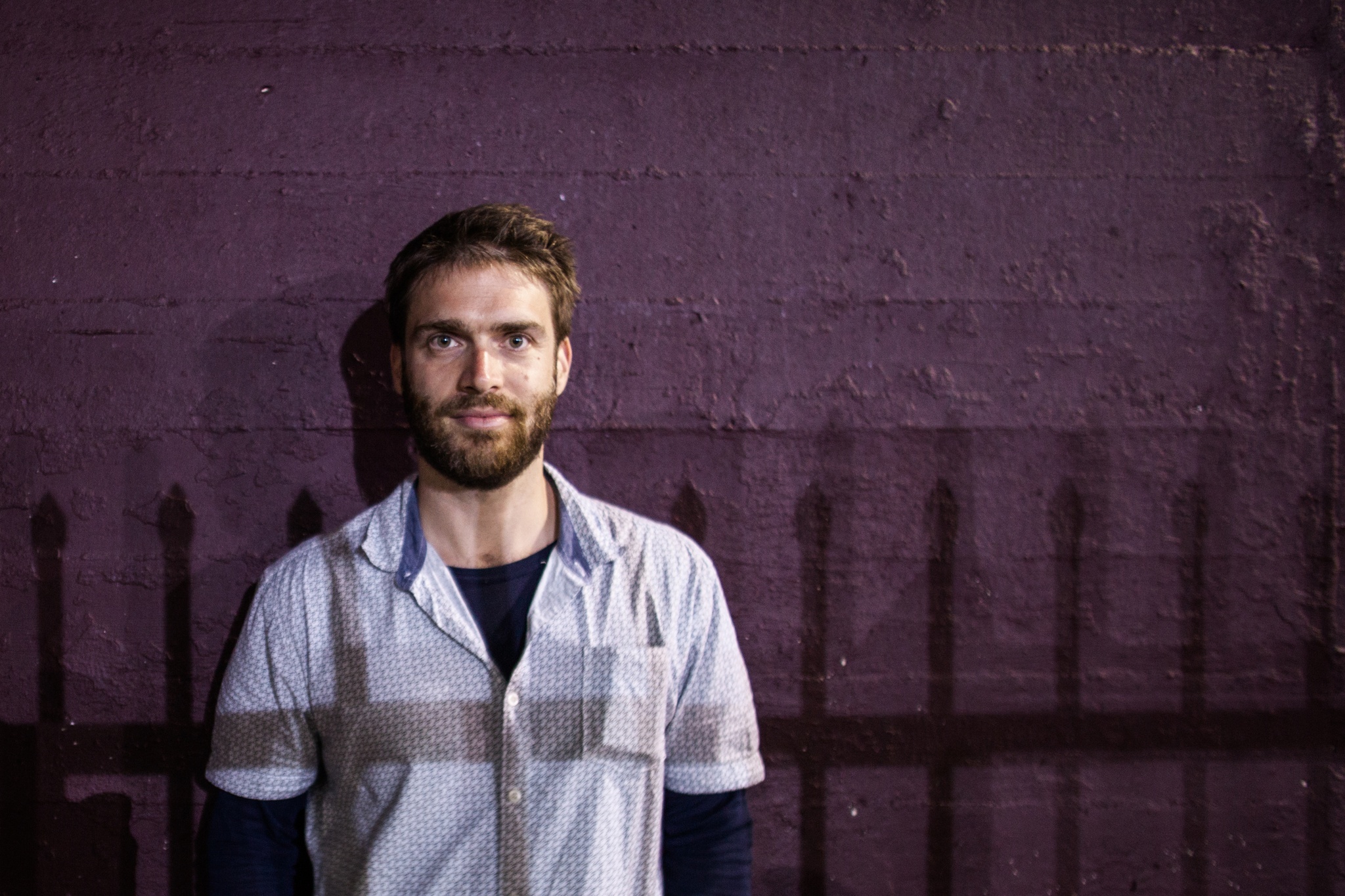 A white man in a light purple button down shirt poses against a dark purple wall. He has short brown hair and a beard and mustache. The shadow of a fence falls across his torso. 