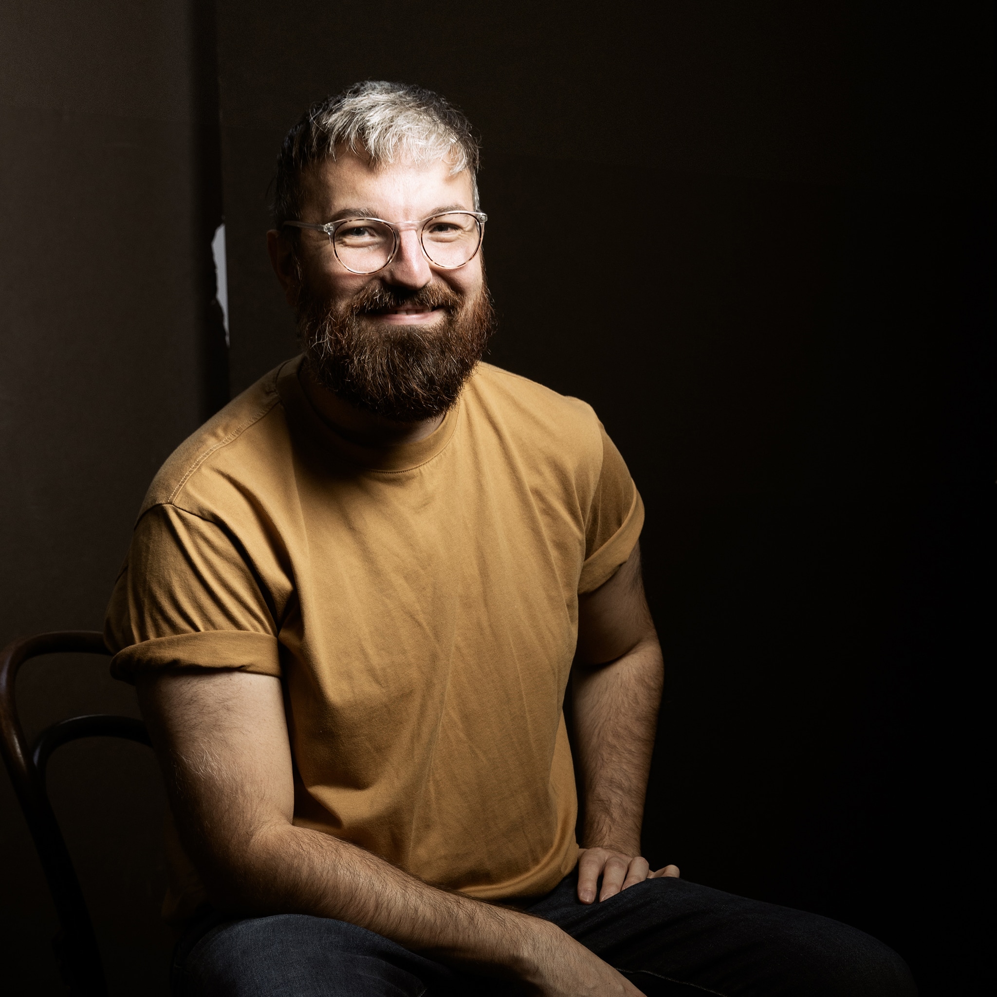 A portrait of artist Andrew Morrill. He is a white man who is pictured in dramatic lighting with the background in shadow. He leans slightly forward on one arm propped on his leg. He had a beard, wears glasses and a golden brown t-shirt, and smiles at us.