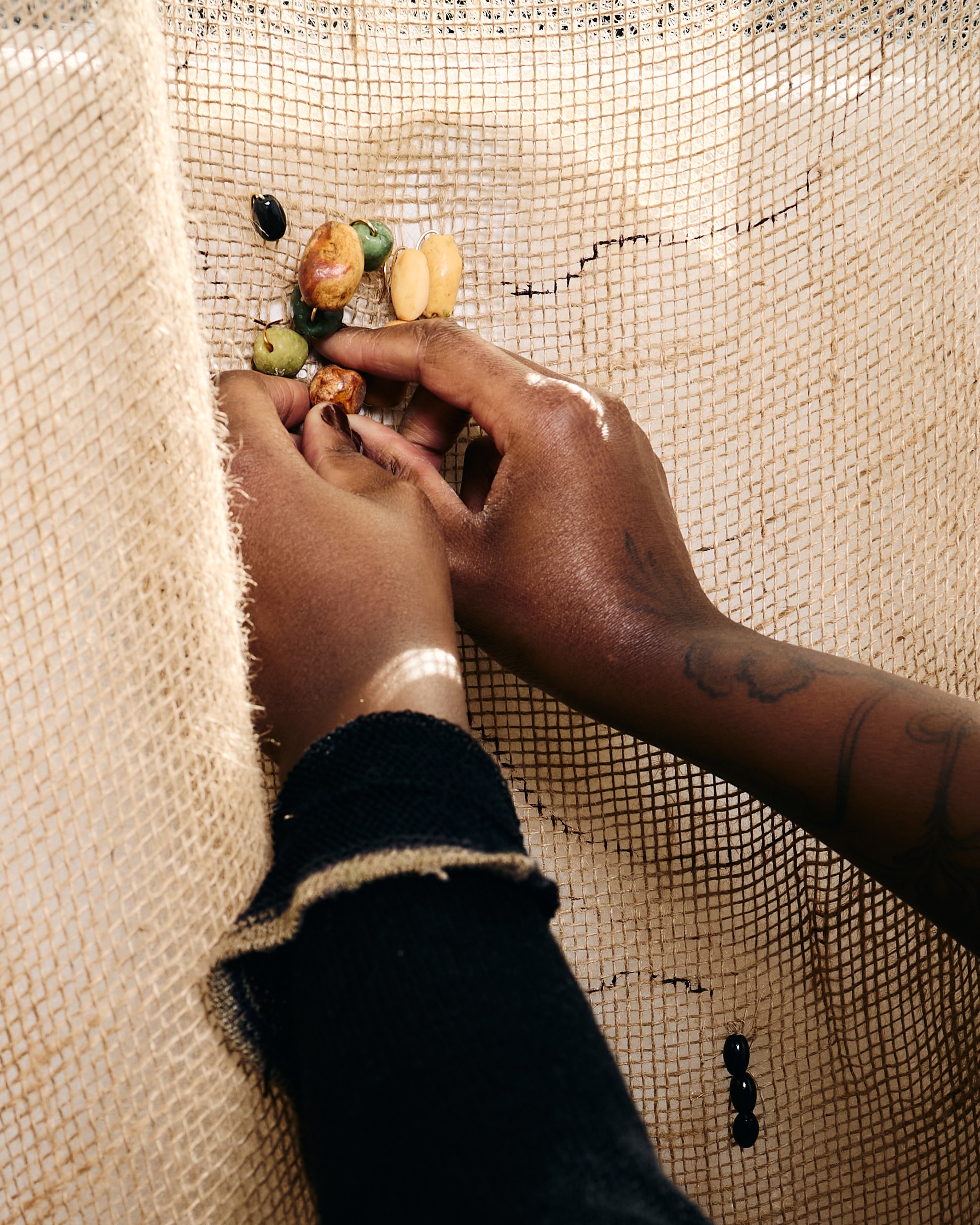 The hands of artist Laurena Finéus are seen in a close-up as she stitches small ceramic sculptures of seeds to a panel of beige fabric hanging in front of her. 