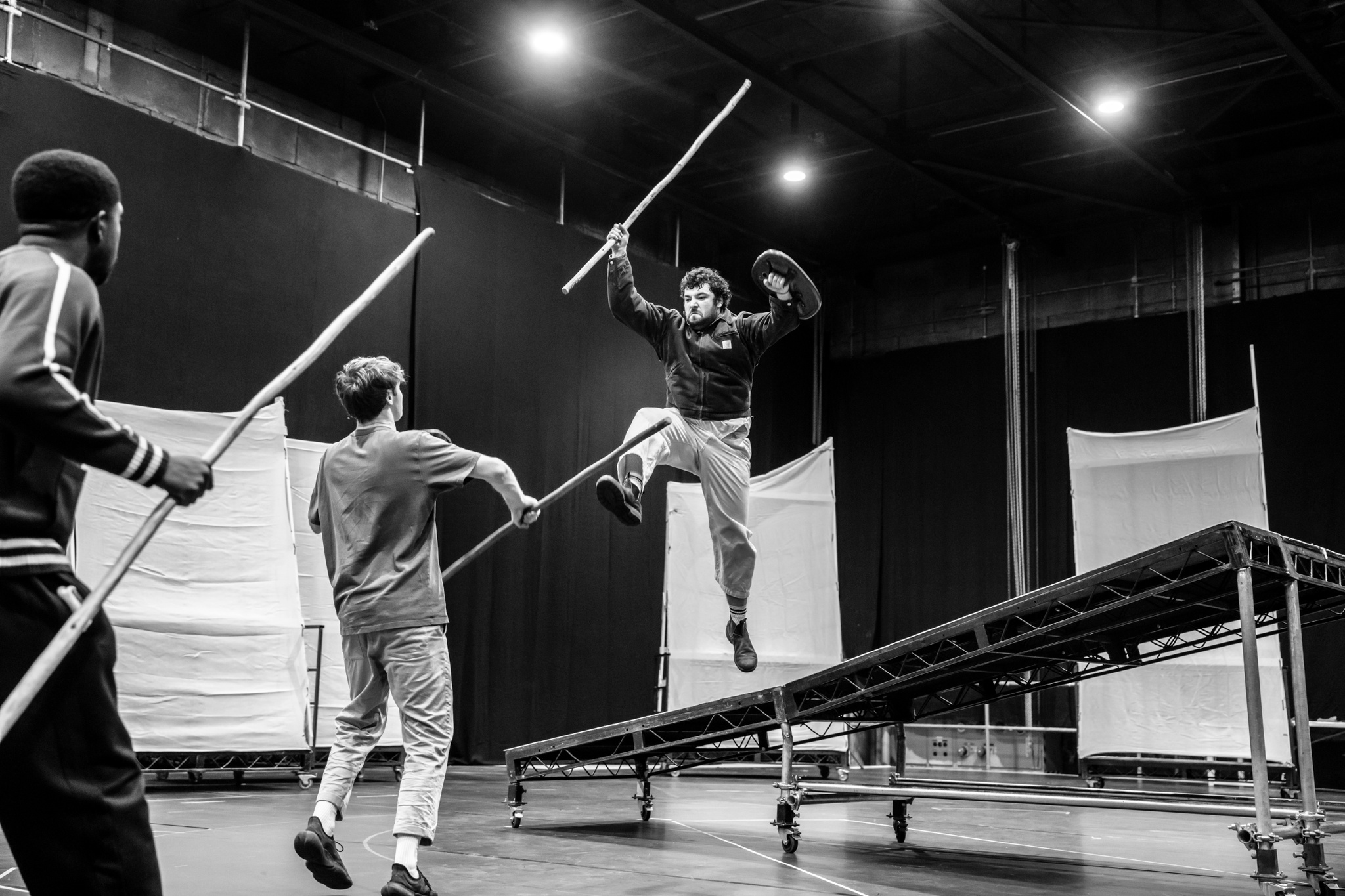 Actors Caleb Obediah, Doug Colling, and Dylan Corbett-Bader rehearse a fight scene. Corbett-Bader jumps from a platform, lunging toward Obediah and Colling who face him on the ground. They each wield a prop staff.  