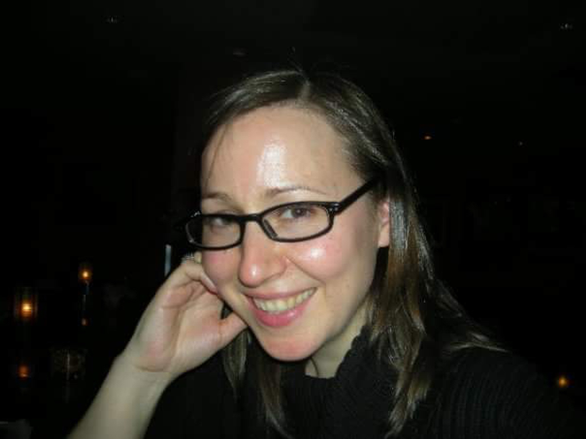 A portrait of Boo Froebel taken in a dark space with a flash so that the background is not clearly visible. Boo smiles and holds one hand to the side of her face. 