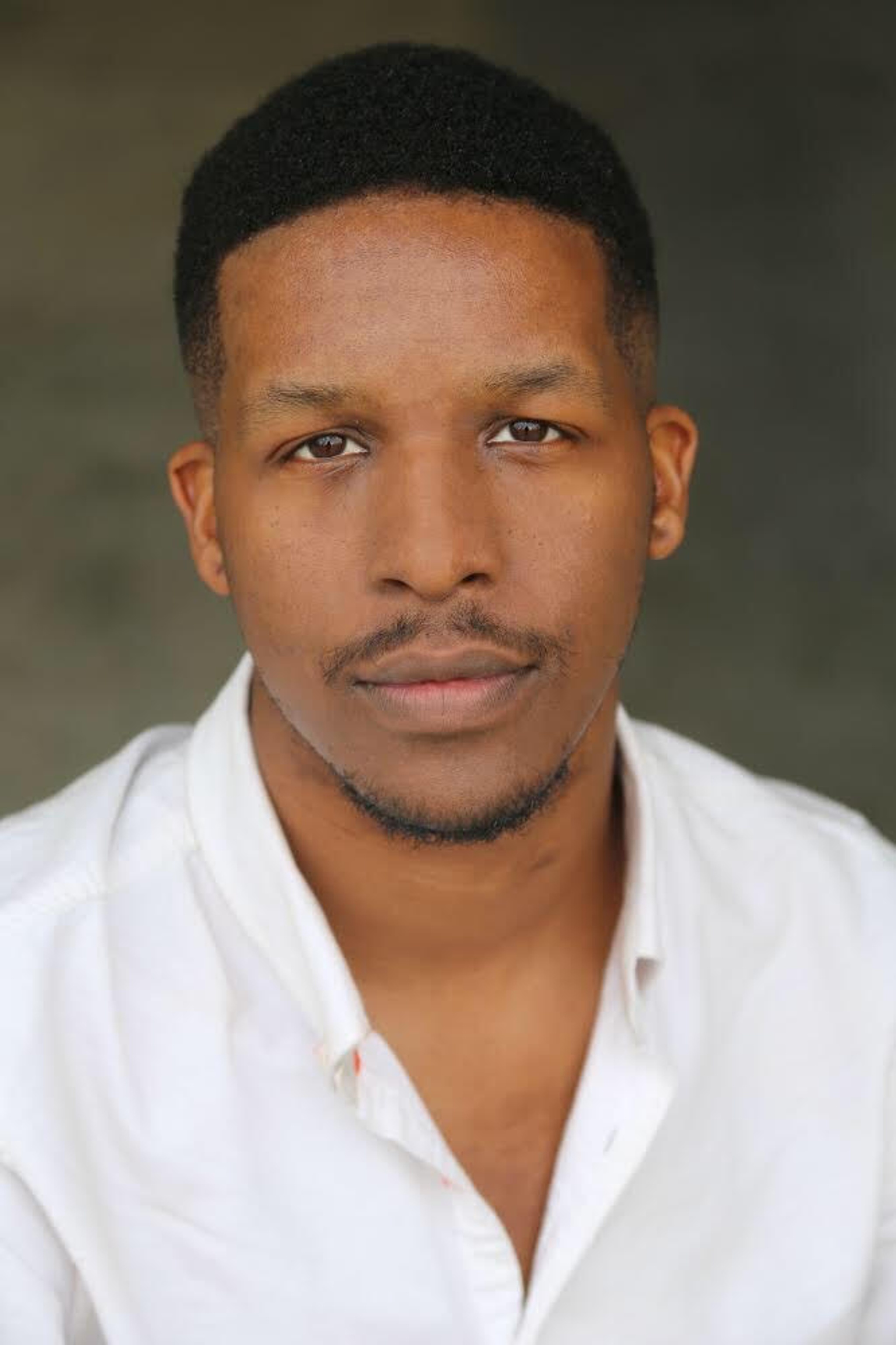 A black man with his hair trimmed in a fade on the sides and a thin mustache and goatee under his chin. He wears a white button-down shirt and looks directly at the camera. 