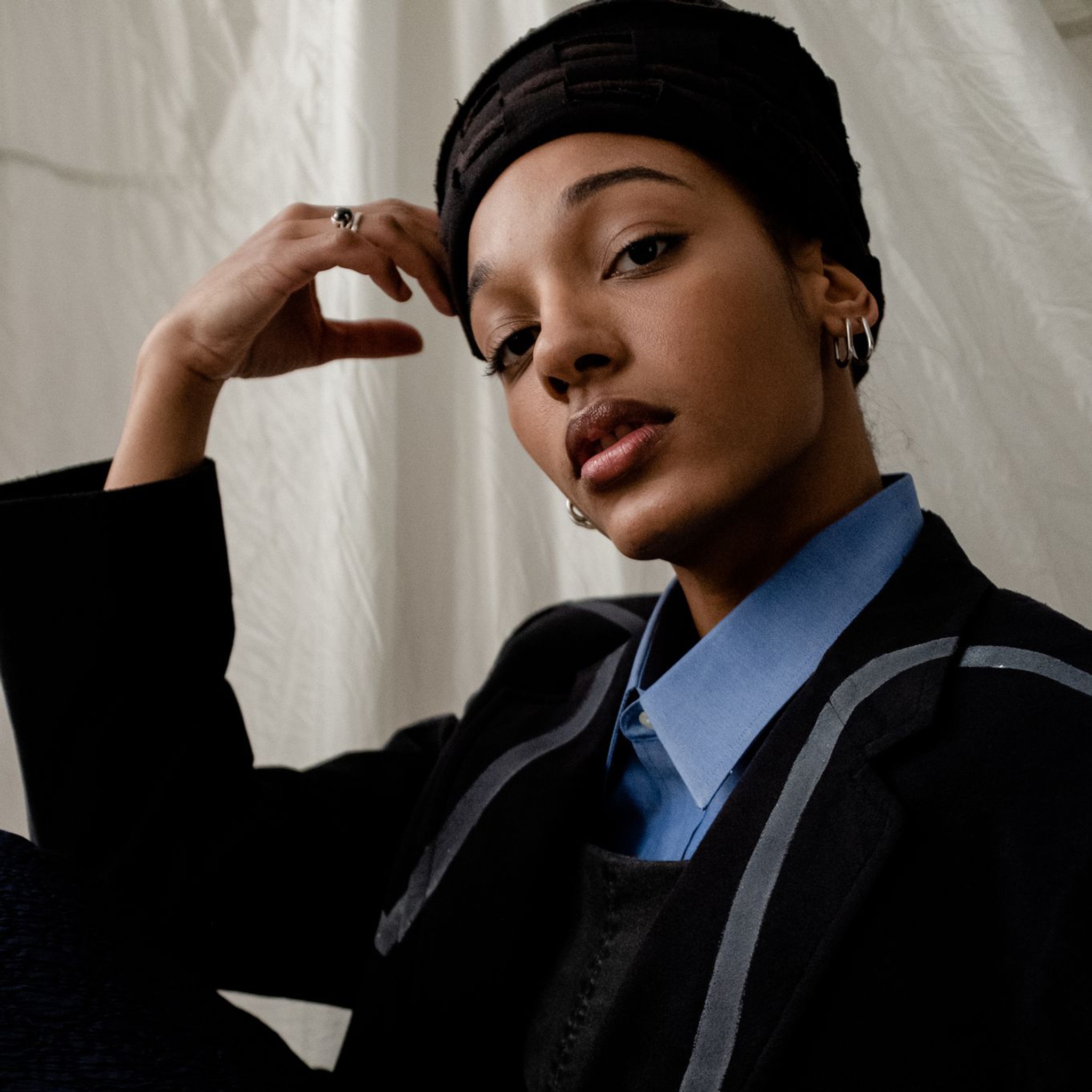 A portrait of Afro-Dominican dancer and choreographer Katherine Paola De La Cruz. Katherine poses in a seated position with one leg drawn to her chest. She rests her elbow on her knee and leans her head one one hand. She wears a jacket over a blue button down blouse. She looks directly at us with eyebrows slightly raised.