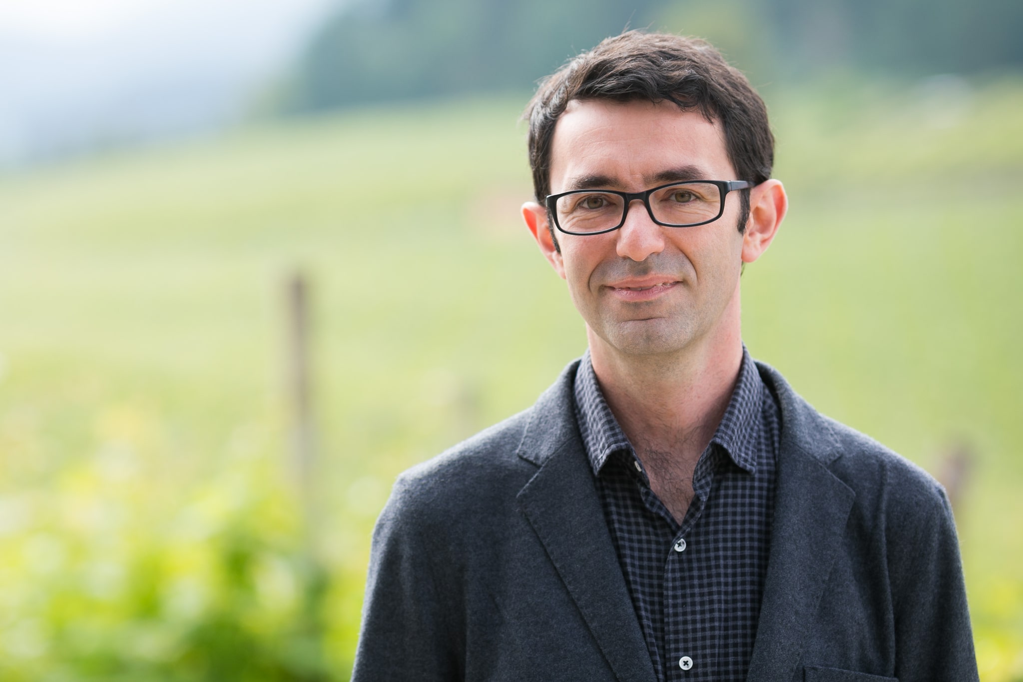 A white man with short dark brown hair and wearing rectangular glasses and a dark gray blazer over a small-print checkered shirt. He's standing with a green rolling field in the background. 