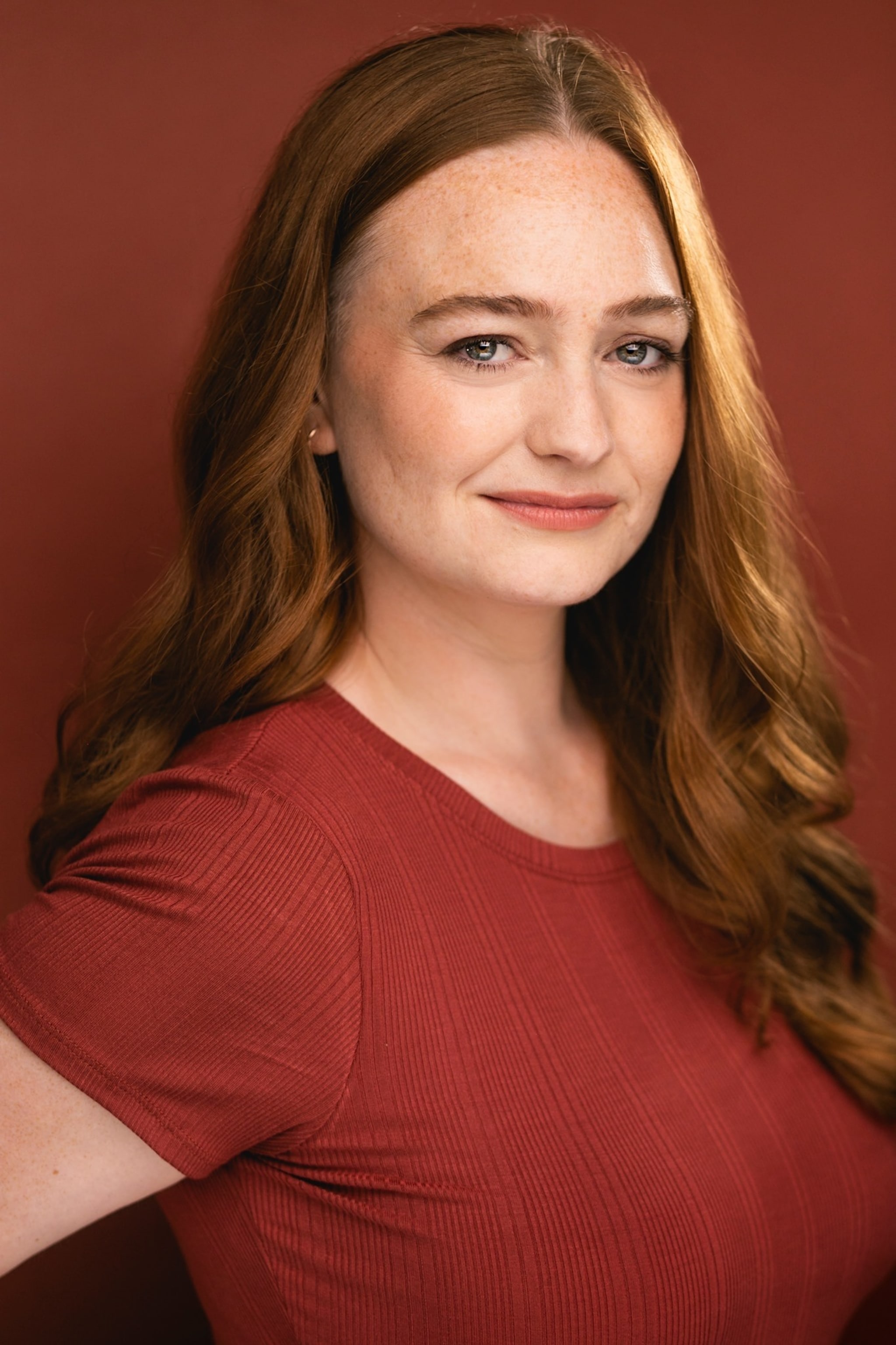A portrait of actor Amelia Hensley, a white Deaf woman with long light brown hair styled in loose curls. She is seen in three quarters view against a terra cotta colored backdrop. She wears a rust colored t-shirt and smiles directly at us. 