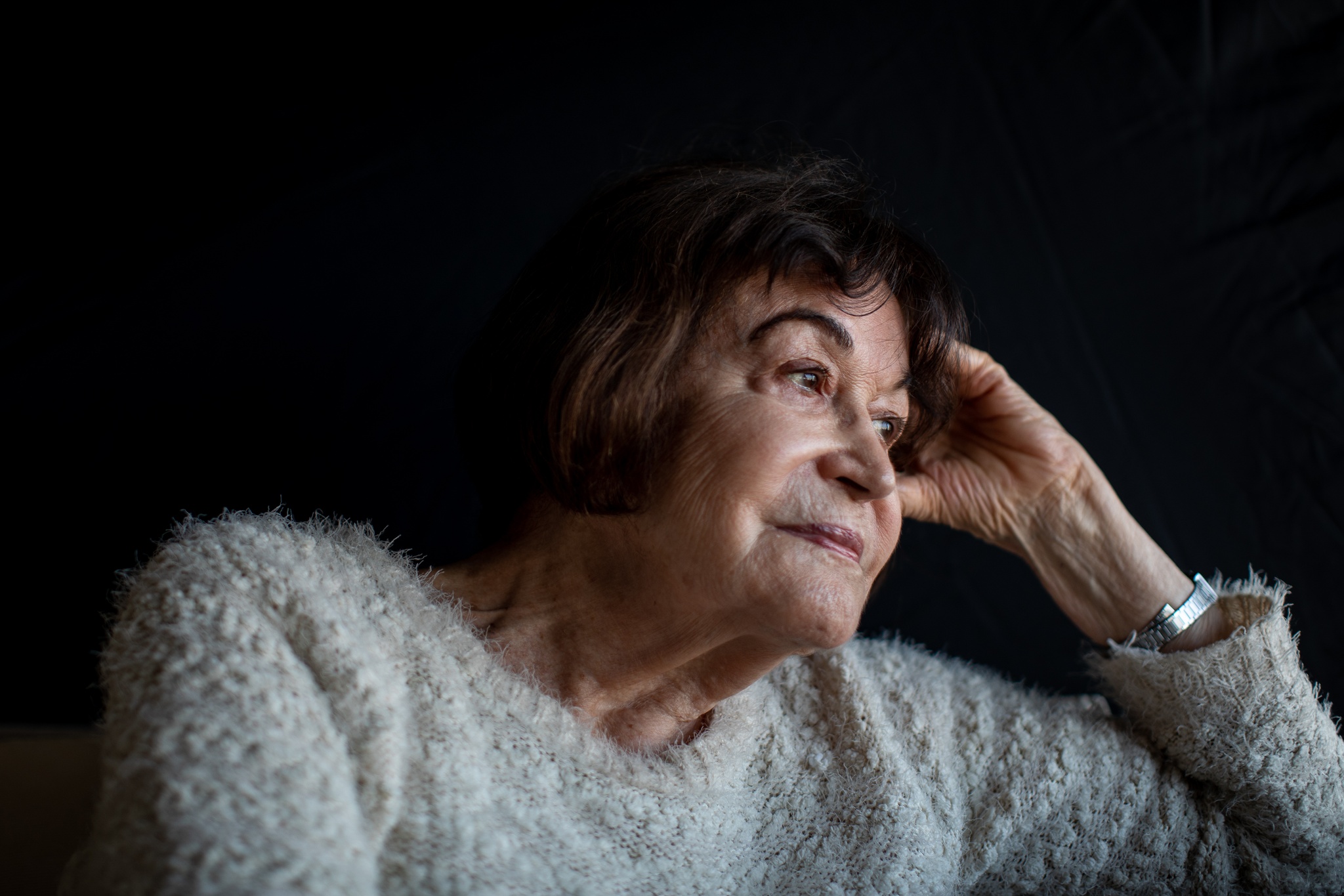 A portrait of artist Claudia Andujar. A white Brazilian woman, Claudia has brown hair in a short bob. She wears a fuzzy sweater and leans her head on one hand, looking away in three quarters view. 