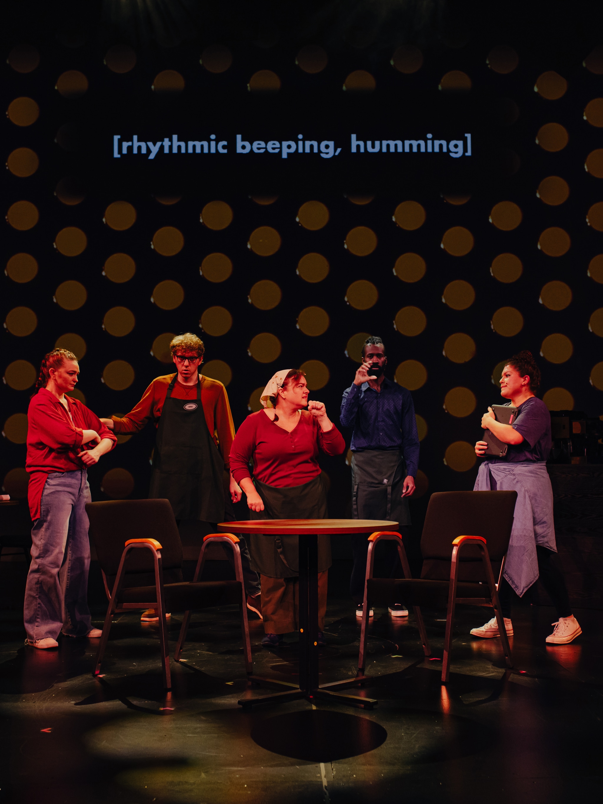 Five Deaf actors stand in a row facing us on a dark stage. In front of them is a cafe table with two chairs. Behind them is a projection of patterned dark orange dots evoking the interior of a microwave. 