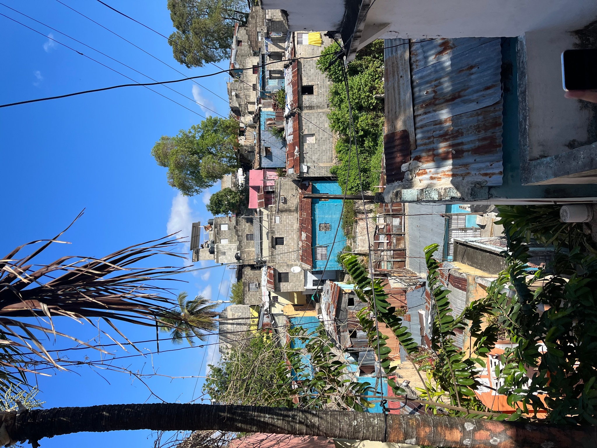 A hillside neighborhood in the Dominican Republic. Houses are stacked up the hill and look as if stacked on top of each other from the perspective of the photographer. They are predominantly a gray cement color but punctuated with some painted light blue and pink. The sky above is a deep blue.