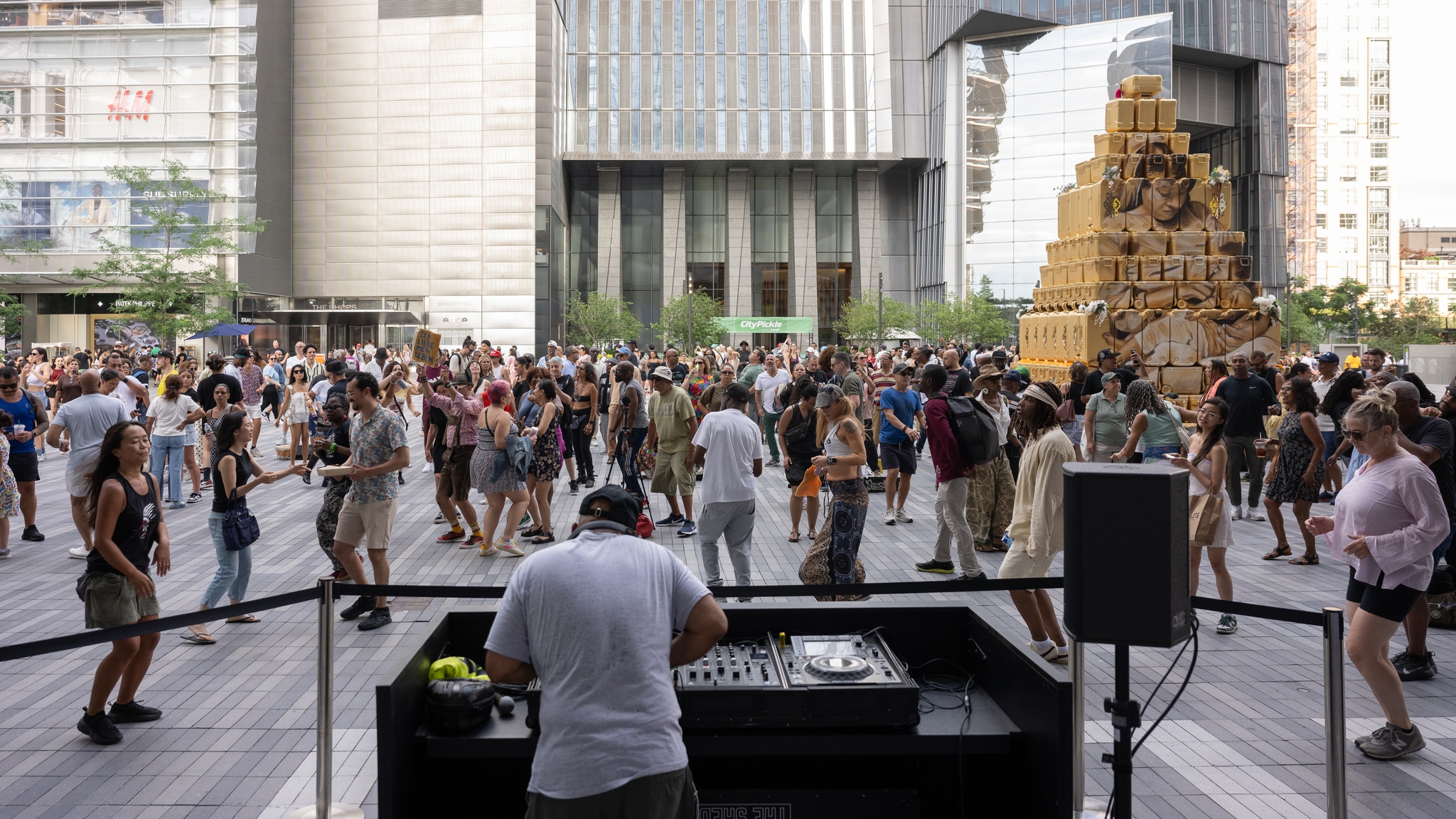 A DJ is seen spinning on an outdoor plaza in front of a dancing crowd