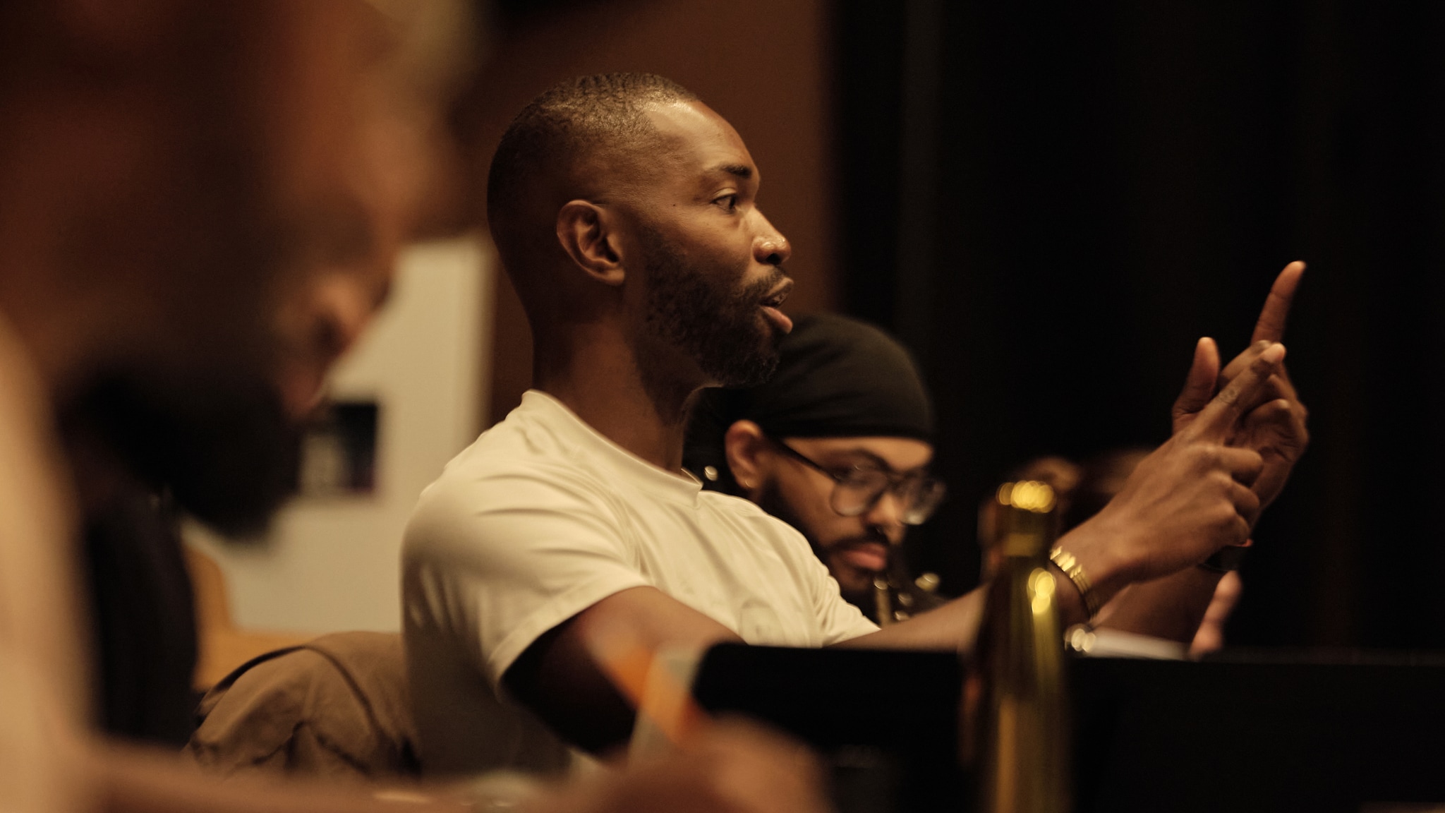 Writer and director Tarell Alvin McCraney, a Black man with a clean shaven head wearing a white t-shirt, is seen in profile from a long angle while he sits in a rehearsal studio. He is gesturing toward someone out of frame, speaking to them with his hands raised. Behind and in front of him sit other members of the production team.