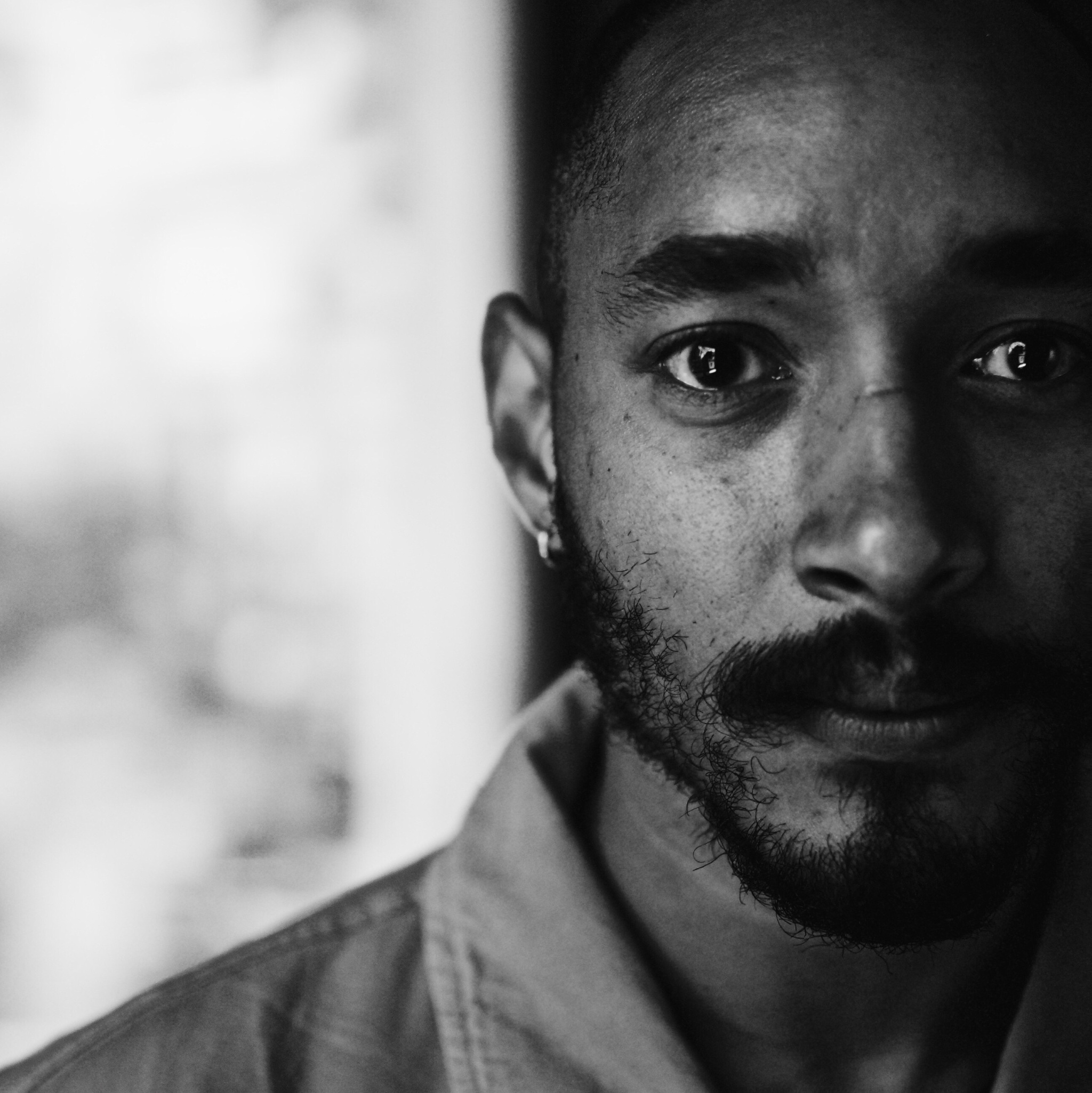 A black-and-white photo of the artist Niall Jones. The photo is a close-up on Niall's face that cuts off the left side of his face. The background is blurry but dappled with light.