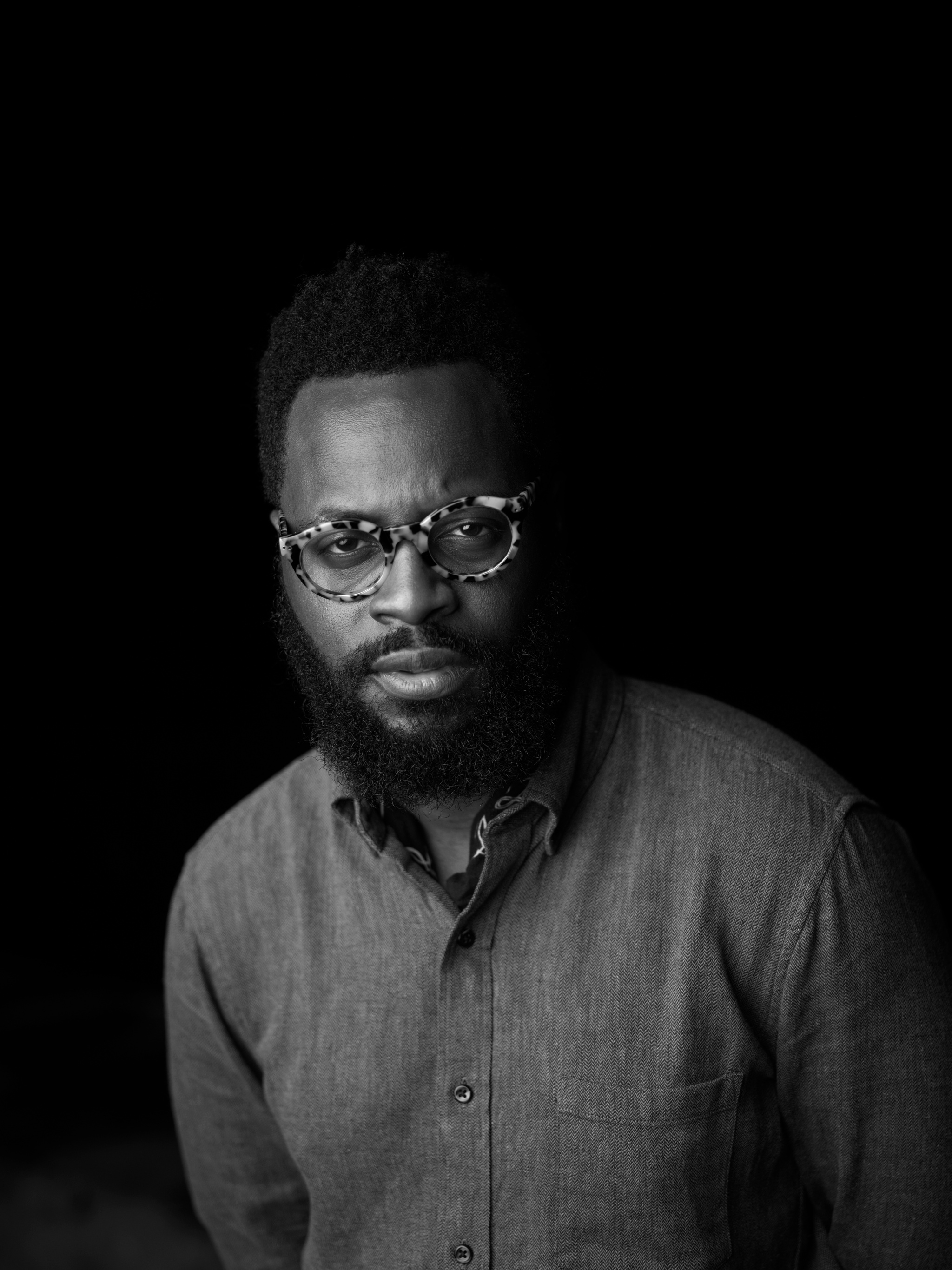 A black-and-white photo of Pythagore Antoine against a black background. Antoine wears heavy, tortoise-rimmed glasses, has a beard and mustache, and has tucked a kerchief tied lossely around his neck beneath the collar of a button-down shirt. 