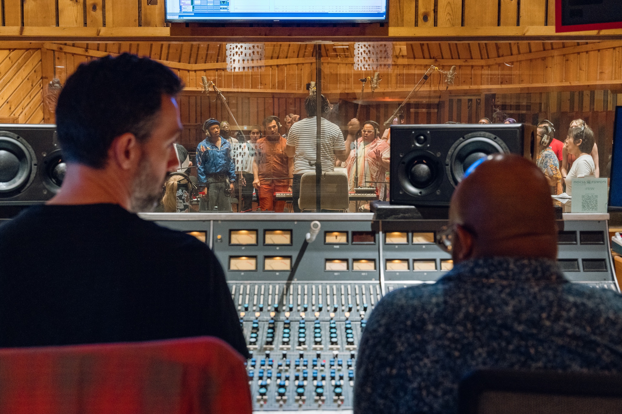 Two men are seen from behind sitting in a recording studio booth. In front of them is a control panel and through a window they look out on a choir in the studio. 