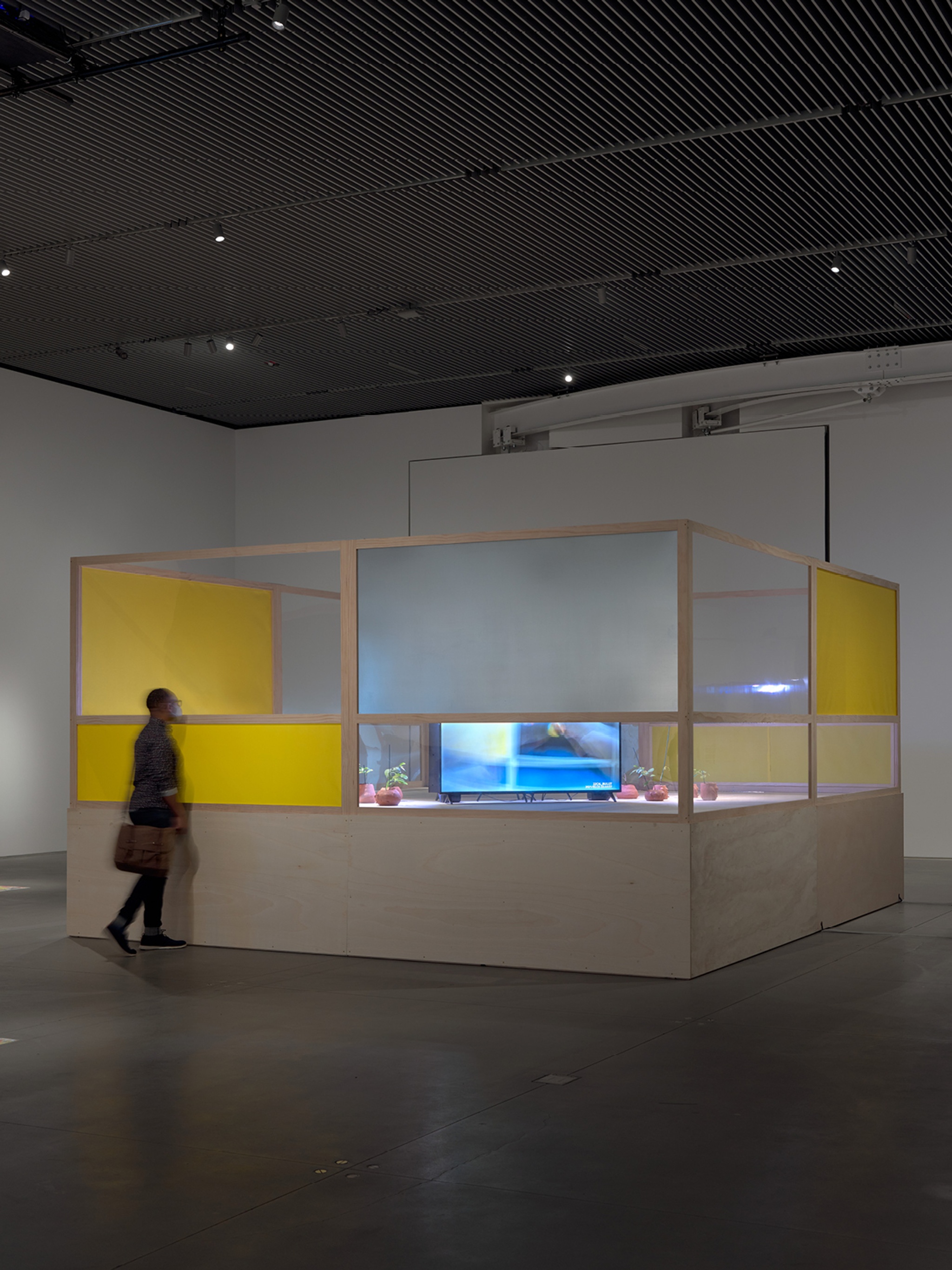 A wooden structure roughly the size and form of a boxing ring sits in an art gallery with a visitor standing in front of it