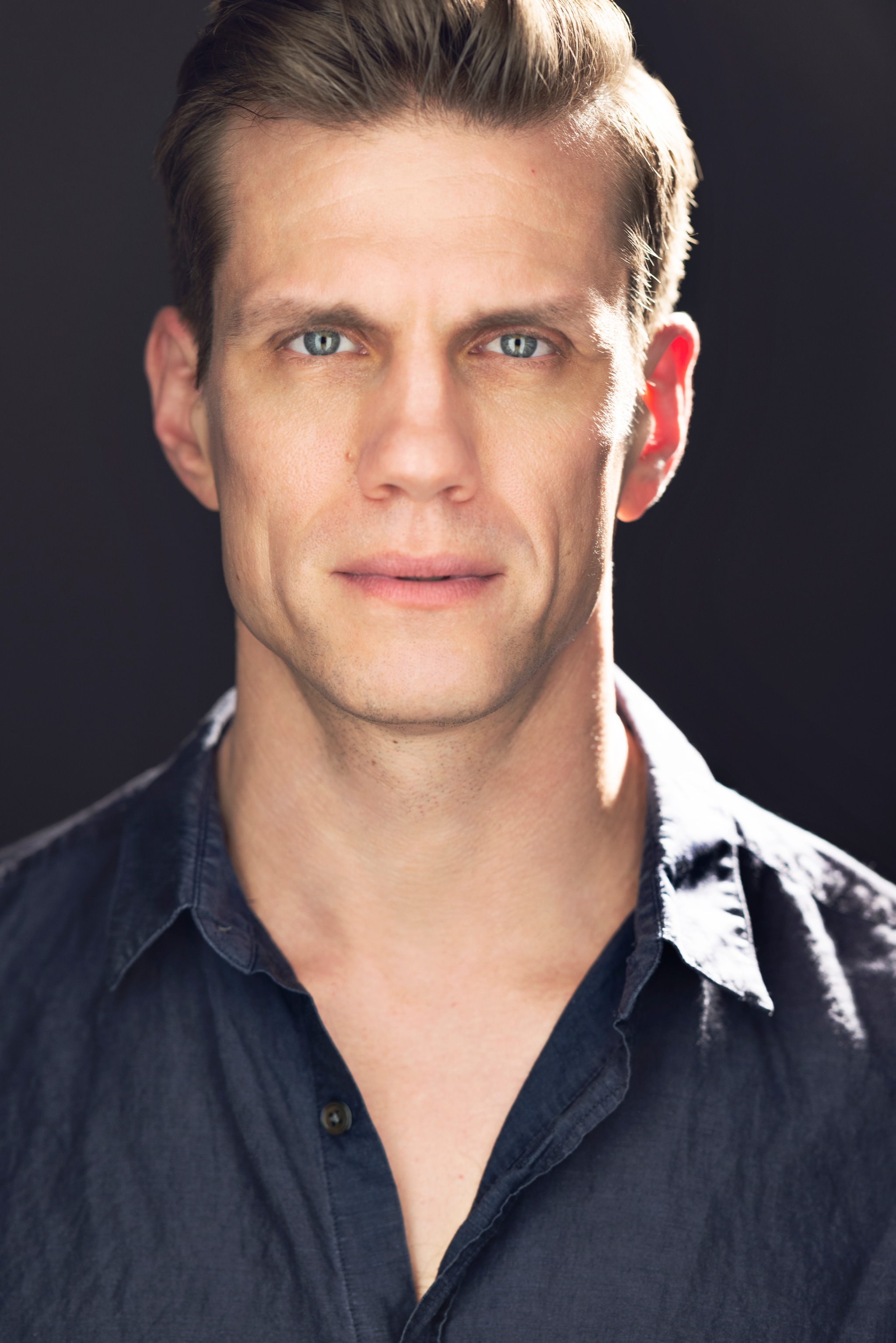 A headshot portrait of Lee Aaron Rosen, a white man with dirty blond hair parted neatly to the left side of his head. He wears a navy button down shirt unbuttoned at the collar and looks intently directly at us. A bright light shines on the left side of his face from the background. 