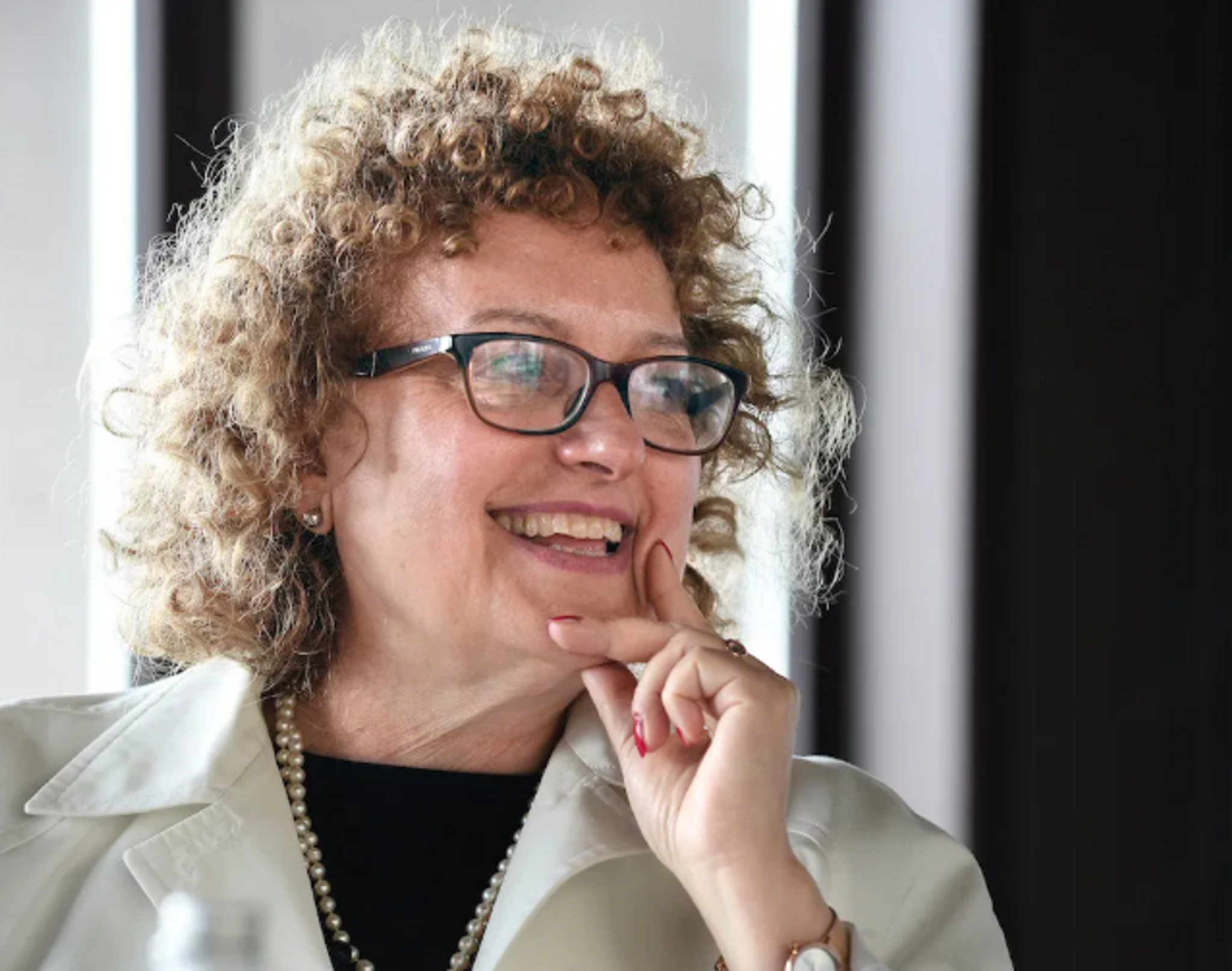 Curator Carolyn Christov-Bakargiev is seen turning slightly to the side laughing with someone outside the frame. She is a white woman with curly light brown hair. She wears glasses and smiles broadly. 