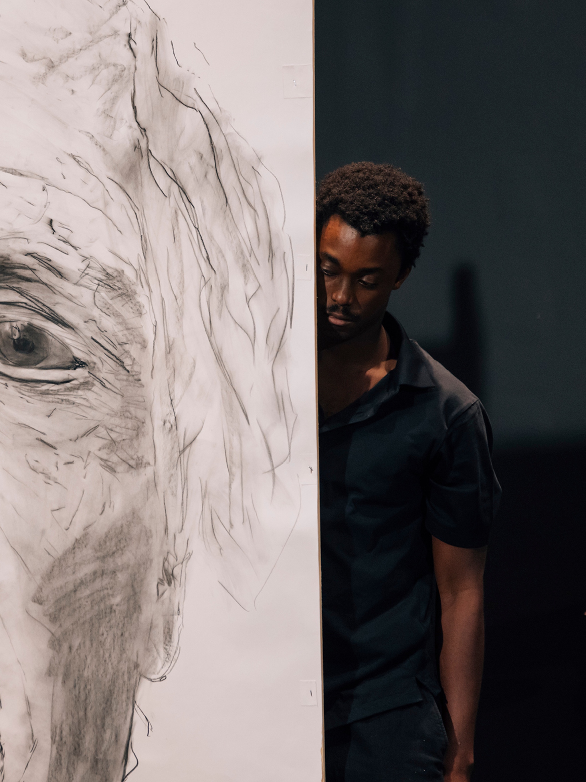 Performing in "minor b", Nile Harris peeks out from behind a tall screen on stage. Nile is a Black man. He wears a black, short sleeved collared shirt, and half of his body is visible and half obscured by the screen. On the screen is a drawing of an older man's face. The photo shows the man's left eye, ear, and a shock of white hair.