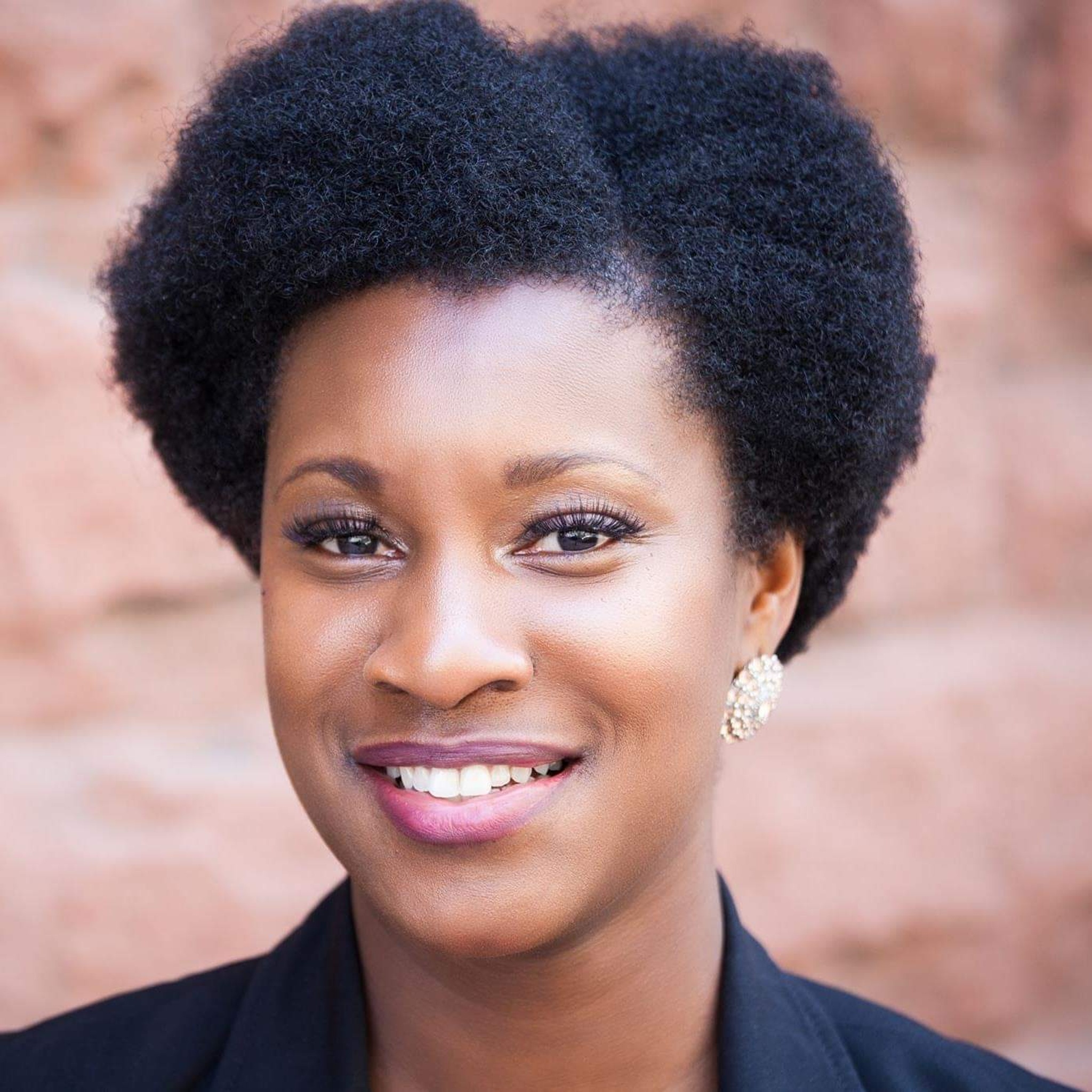 A close-up portrait of Xamayla Rose, smiling with her hair part in a diagonal along he left side of her head in front of a blurry red-brick background. 