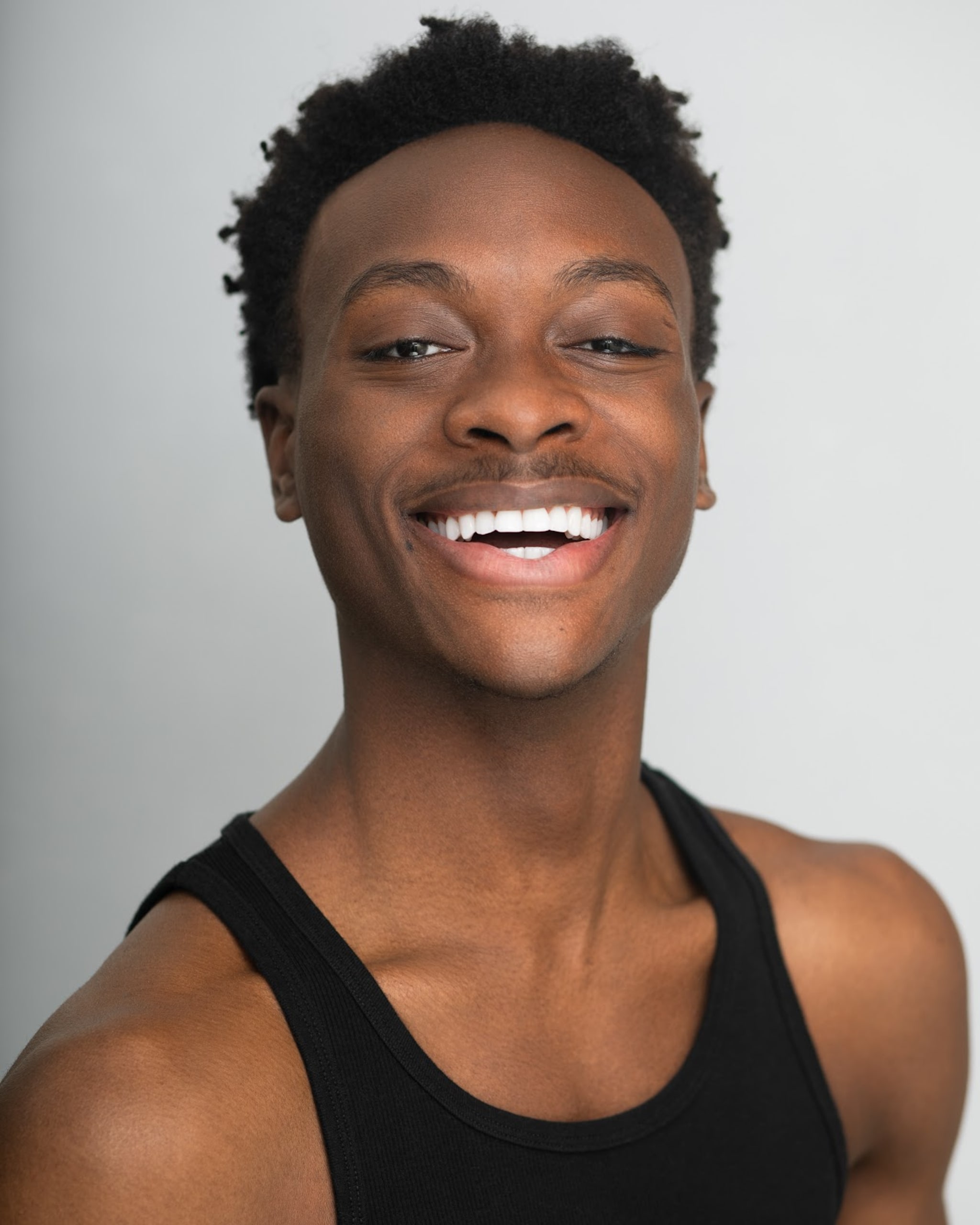 A portrait of performer Kamau Nosakhere, a Black Barbadian person with short dark brown hair. Kamau wears a black tank top and tilts his head back slightly as he smiles broadly at us. 