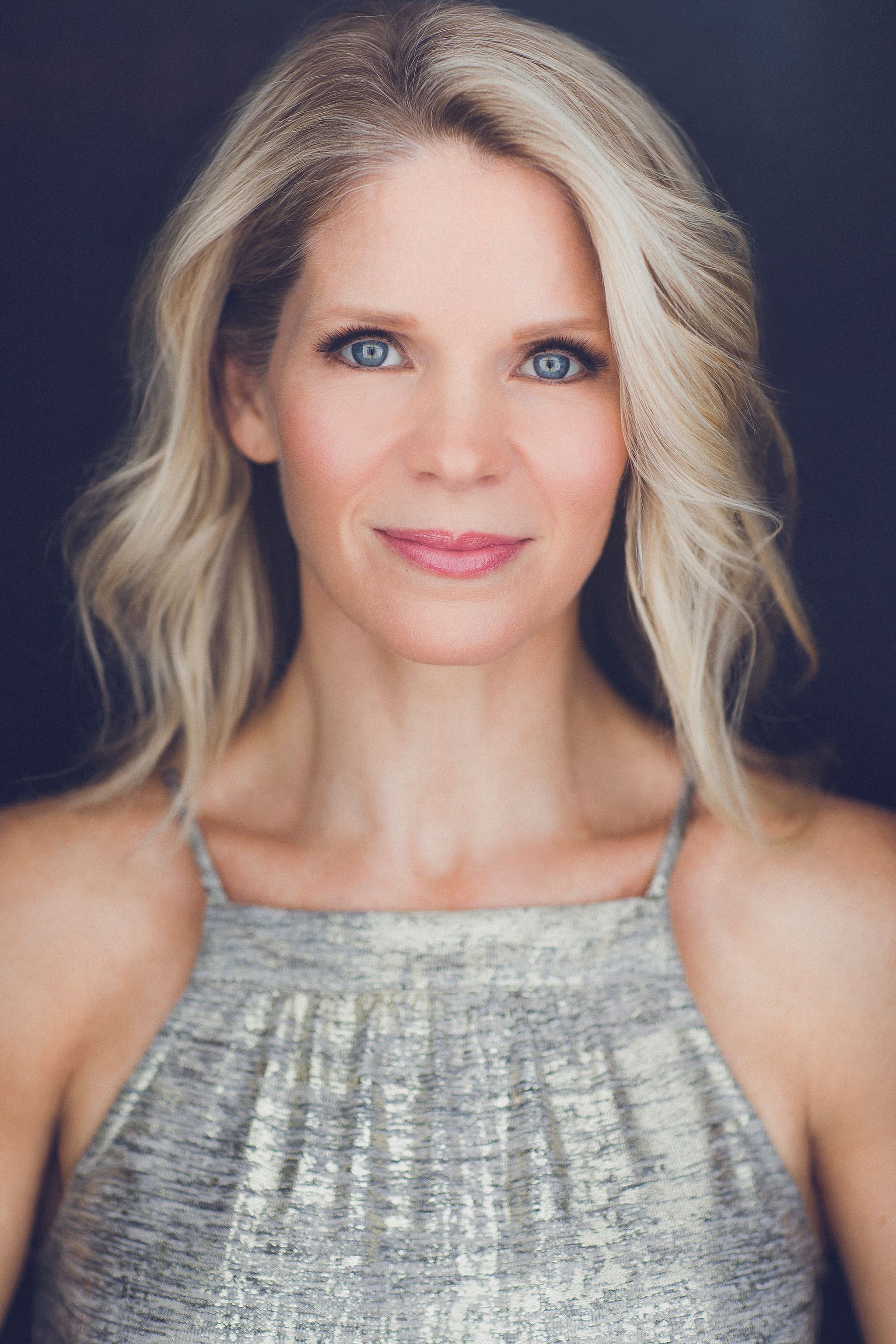 A portrait headshot of actor Kelli O'Hara. Kelli is a white woman with blond, shoulder-length hair parted on the side. She poses against a dark background, looking directly at us with a slight smile on her face. She wears a shimmery silver sleeveless top.