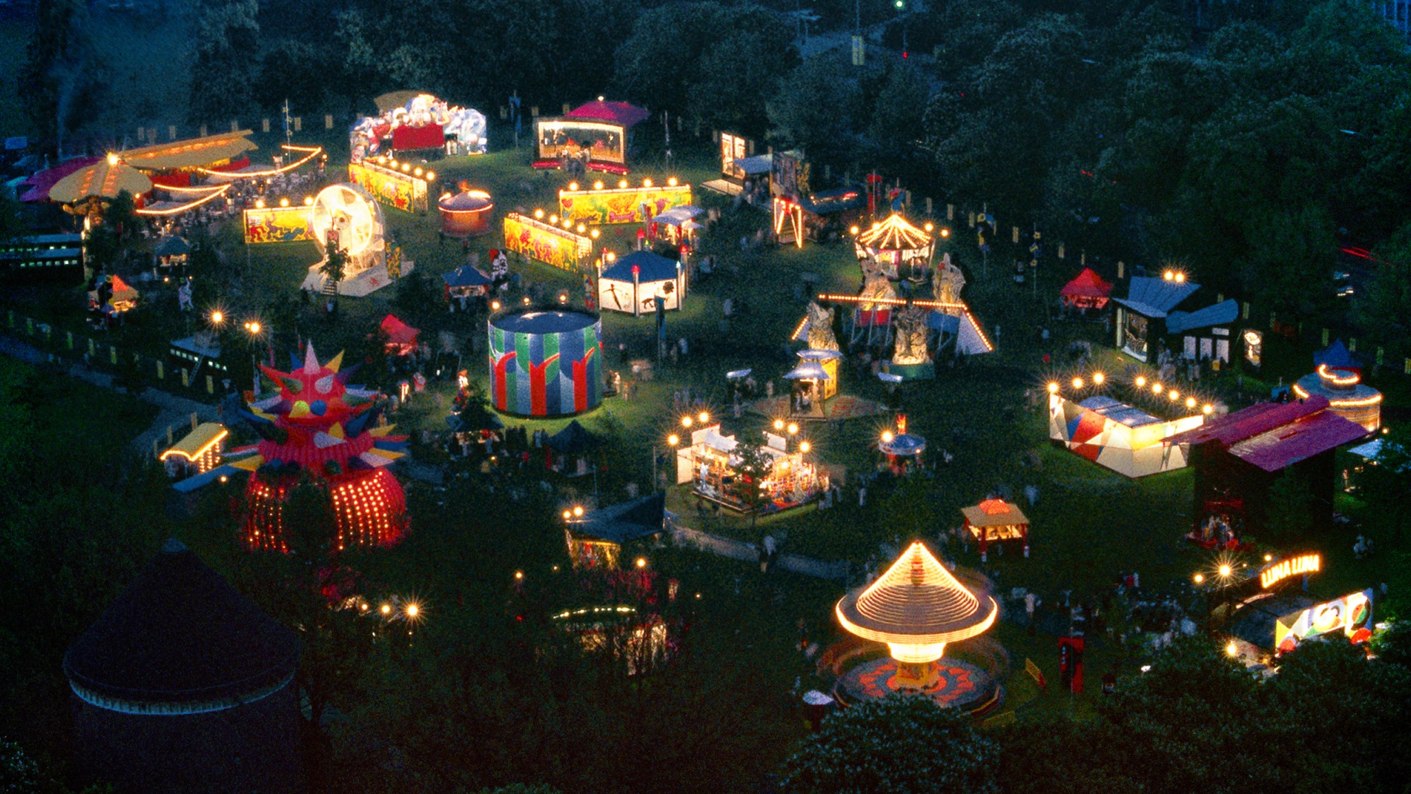 An aerial nighttime view of the original Luna Luna carnival in Hamburg, Germany, 1987. The carnival rides are illuminated by bright light bulbs, giving a festive feeling. 